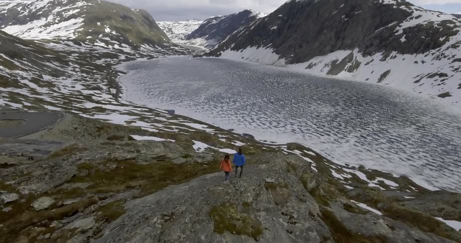 Two people walk to a ledge overlooking a snowy lake and mountains