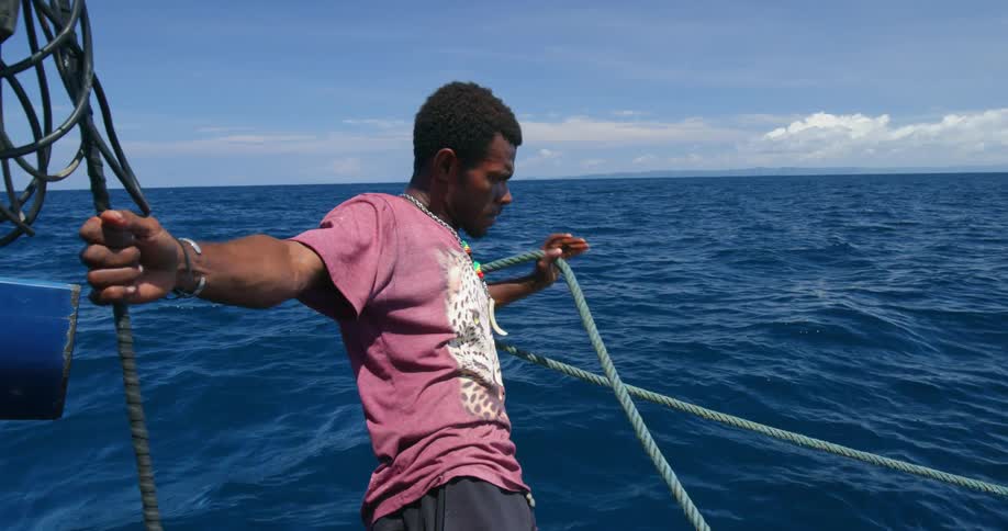 A man leans off a boat on the ocean while hanging onto a cable with one hand and pulling a rope with the other