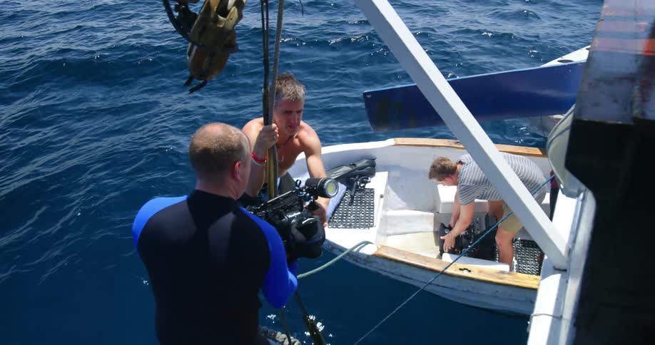 A man passes a large water camera to another person who places it in a small boat