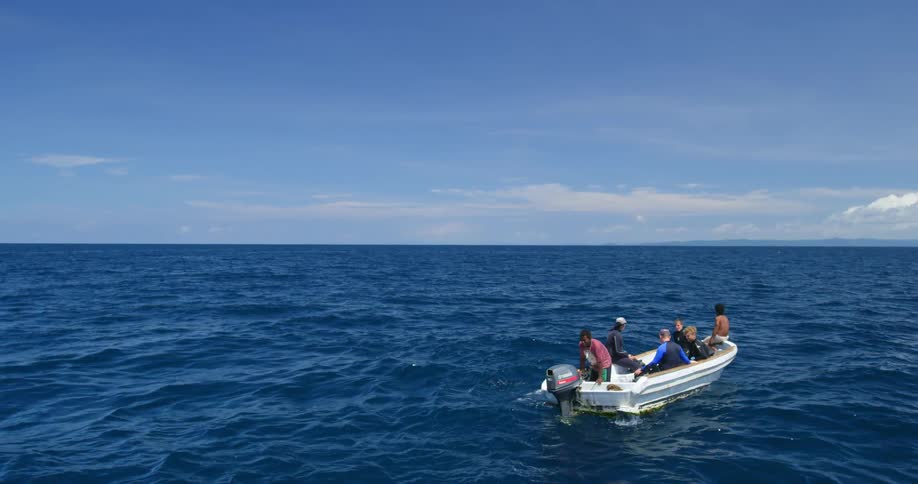 A group of men sit on a boat on the ocean as one man attempts to start the motor