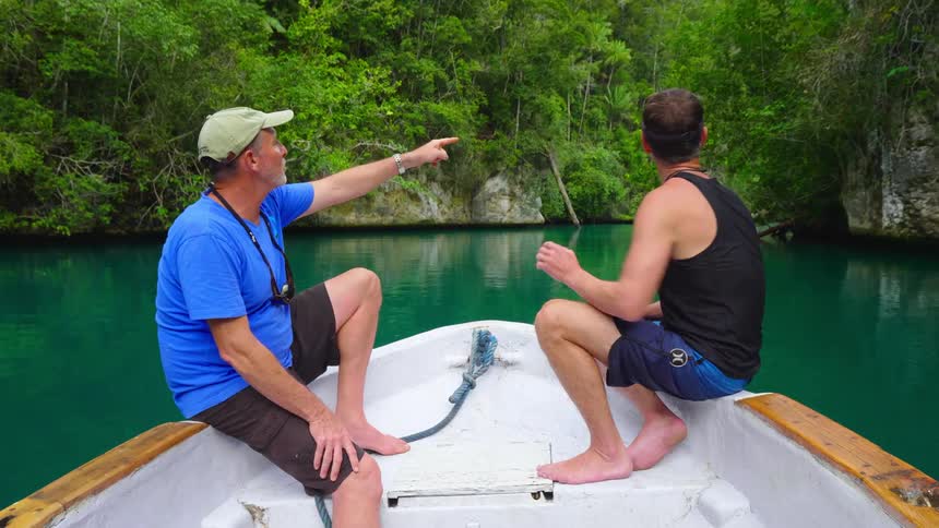 Two men sit on a boat and look at the island they are sailing towards