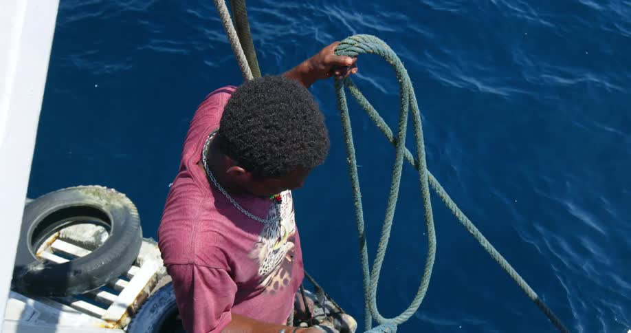 A man on a boat unravels a blue rope thats tied to a surfboard that another person is standing on in the ocean