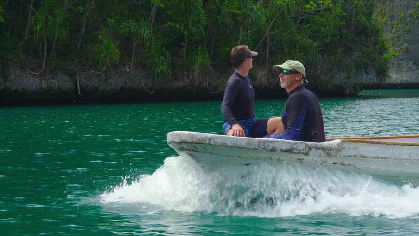 A pair of graying men in wet suits sit at the front of a small boat as it travels between islets on the ocean
