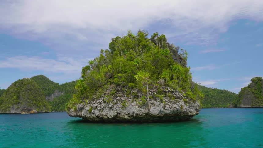 An small rocky island is surrounded by water in a tropical bay area with many hills covered with trees