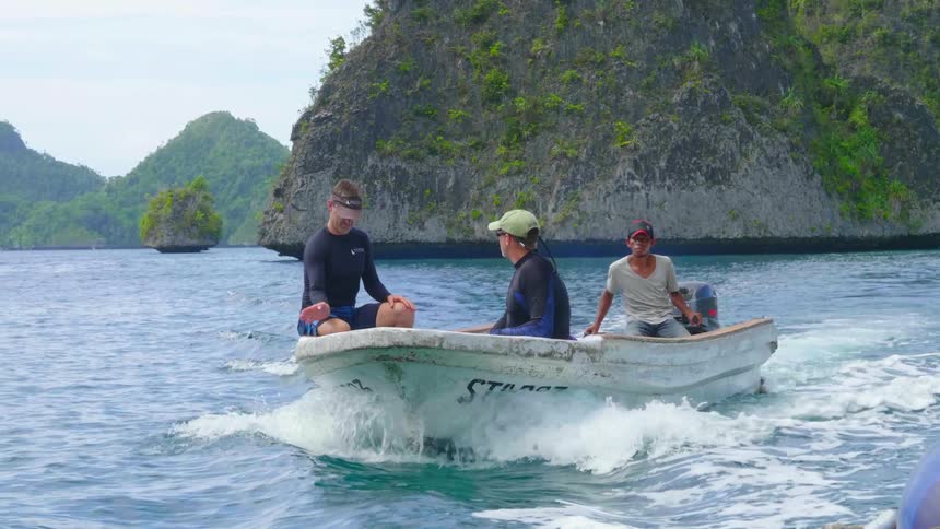 A pair of men in wet suits sit at the front of a small boat as a third man pilots the boat through the ocean