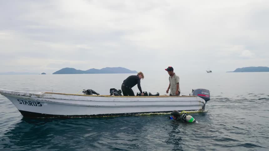 A pair of divers dive off a boat and wait for the people on the boat to hand them underwater filming equipment