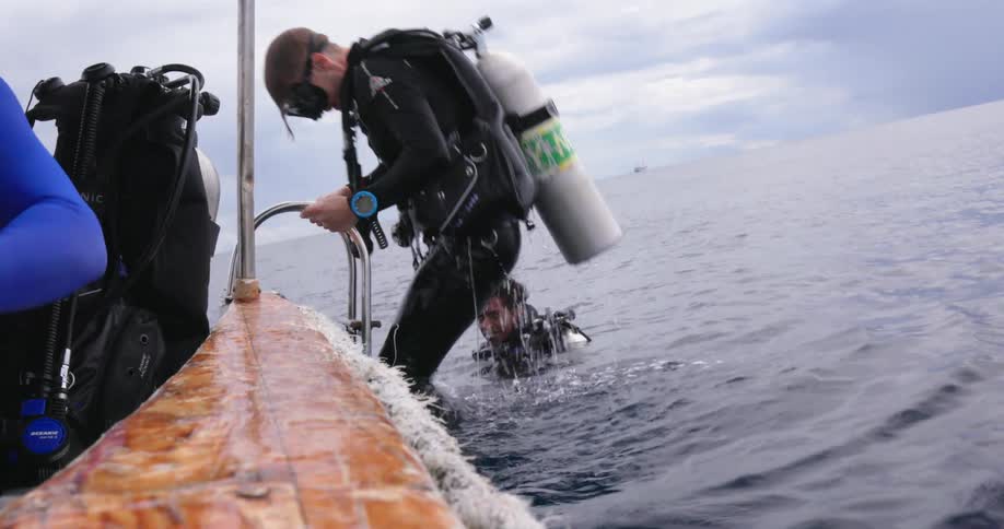 A man with an oxygen tank goggles and a scuba diver suit climbing up a ladder into a boat with the sky and clouds in the background