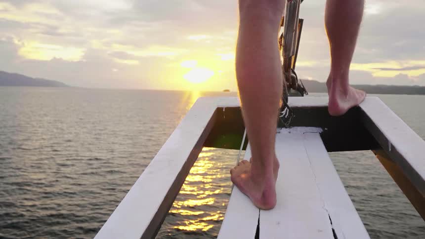 A man stands barefoot on the bowsprit of a boat as it sails through the ocean