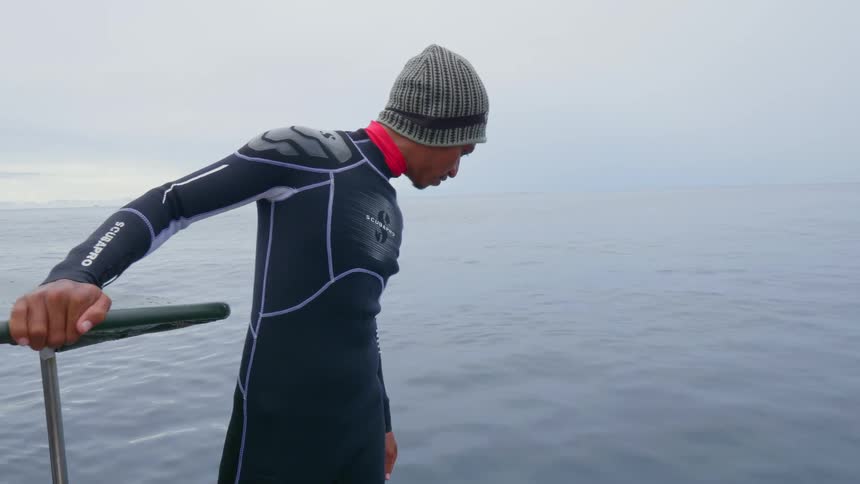 A man in a wetsuit scans the ocean while standing on a boat on a cloudy day