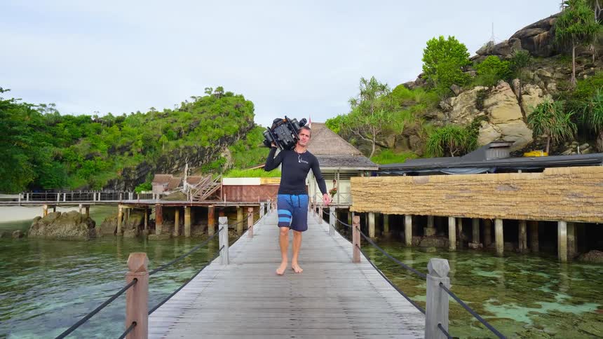 Man carries machine along wooden bridge over green water past buildings and away from forest covered slope