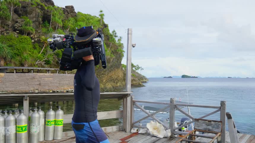 A man carries a large under water camera along a dock towards a boat