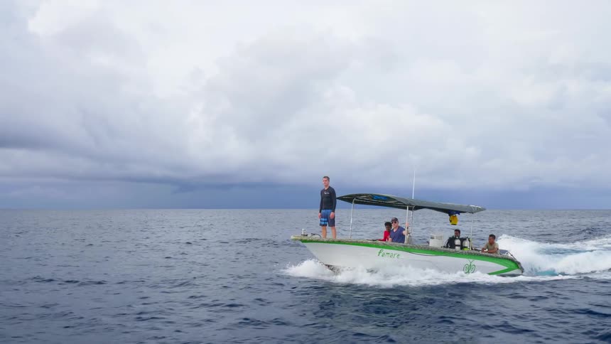 A man stands at the front of a boat as it carries a group of people across the ocean on a cloudy day