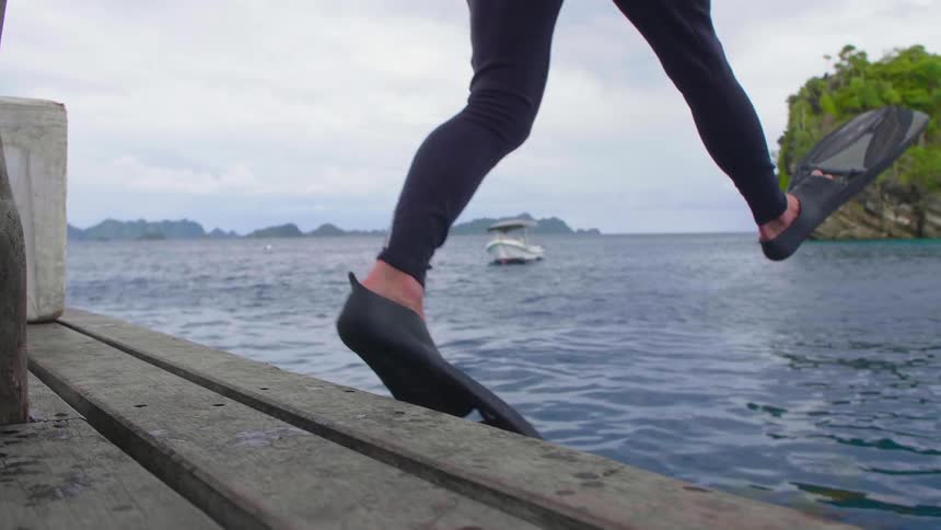 A snorkeler in a wetsuit steps off a pier into the ocean on a cloudy day