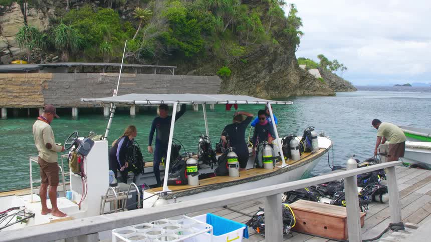 A group of people standing on a docked boat filled with diving equipment adjust their wetsuits