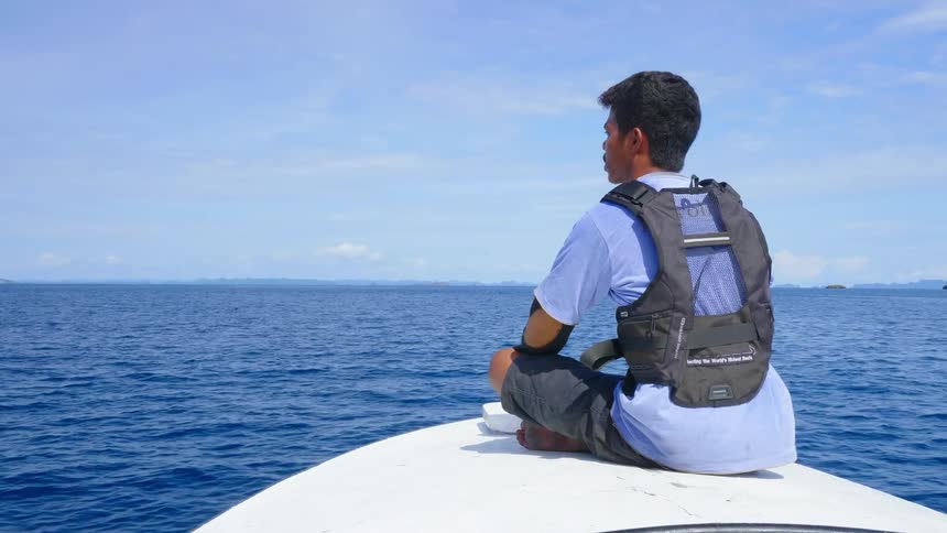 A man wears a black safety vest while sitting on the bow of a boat sailing through the ocean