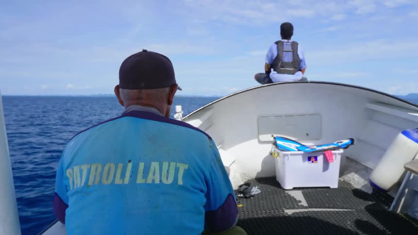 One man sits in boat while another sits on prow looking over ocean toward horizon