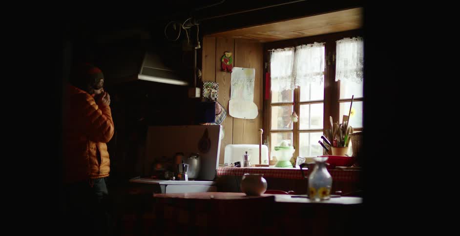 A man stands at home with his hands in front of his mouth and looks out the window