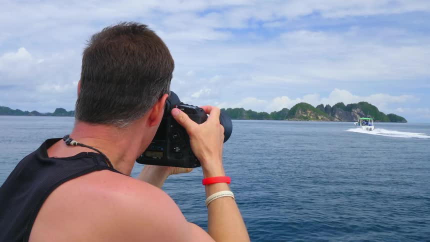 A man on a boat holds a camera up taking photos of another boat on the lake