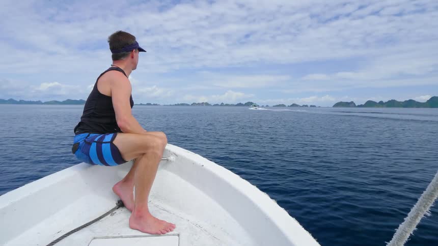 Man sits in prow of boat cutting through ocean toward shore under cloudy sky