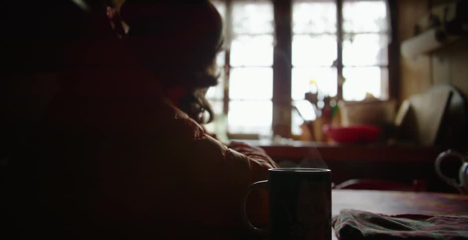Person wearing jacket and cap sits in booth with steaming coffee mug staring toward window across another table