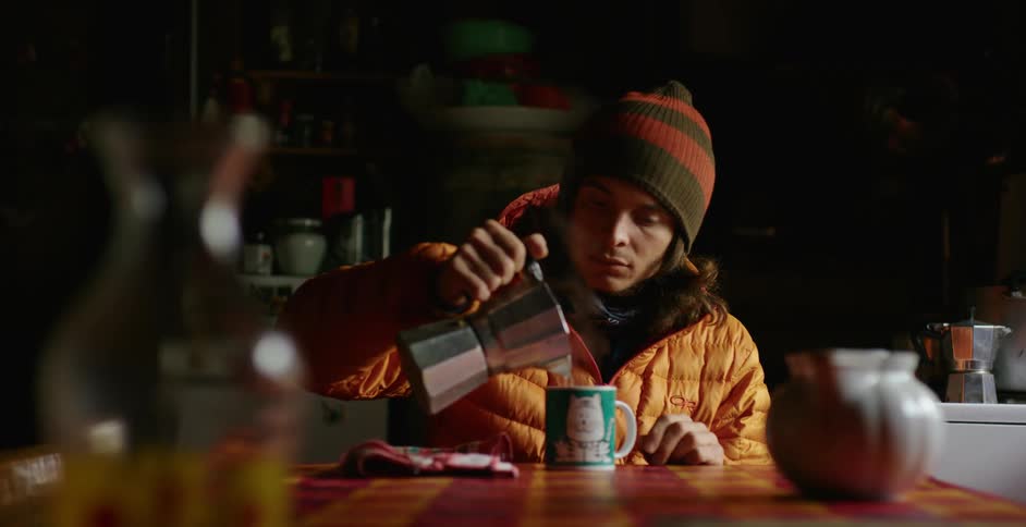 A man pours hot water from a tea pot to a mug while wearing a large coat and beanie
