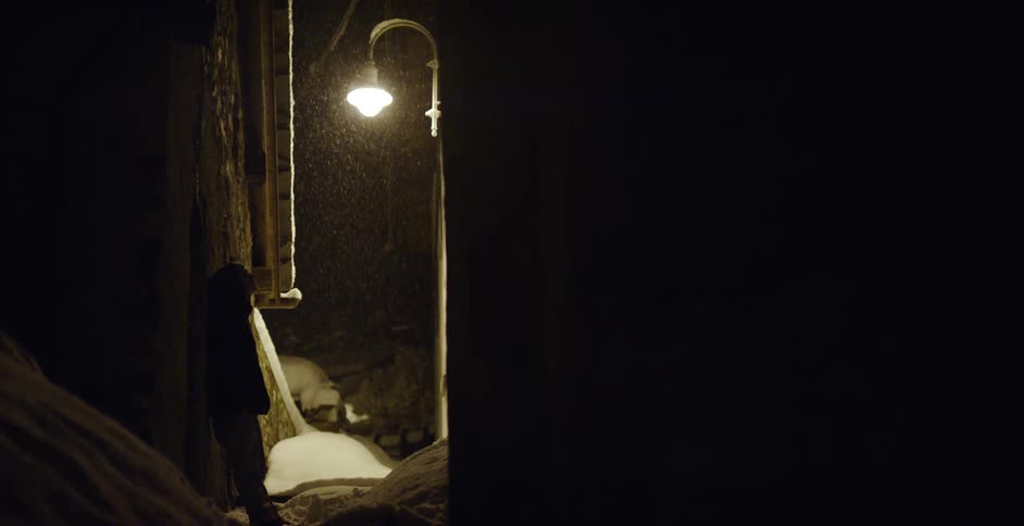 A person leaning against the house in the dark alley as it snows