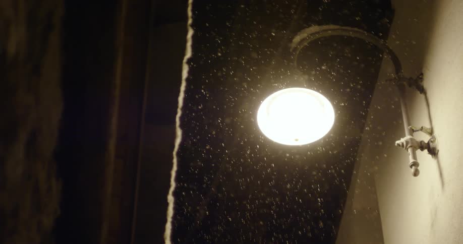 Snow falls around a light hanging on the outside of a building at night