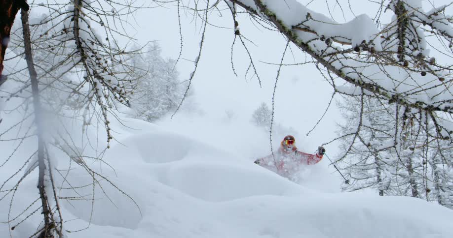 A person with a hat goggles and gloves skiing down a snowy slope with trees