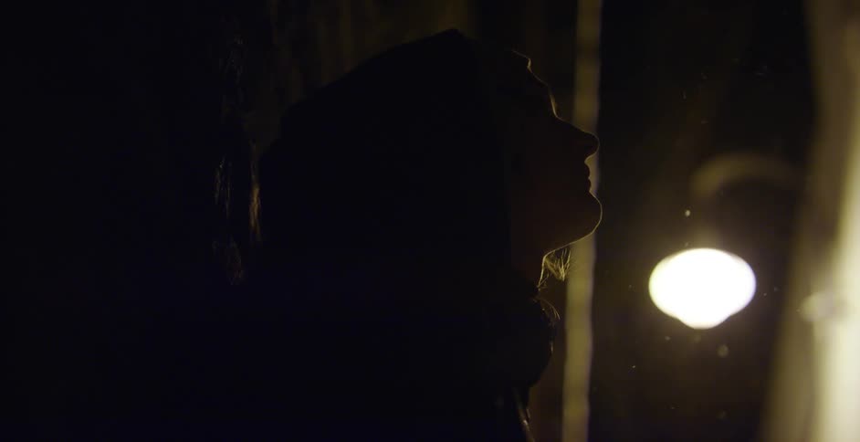 A person stands outside in the shadows next to a building with a bright light