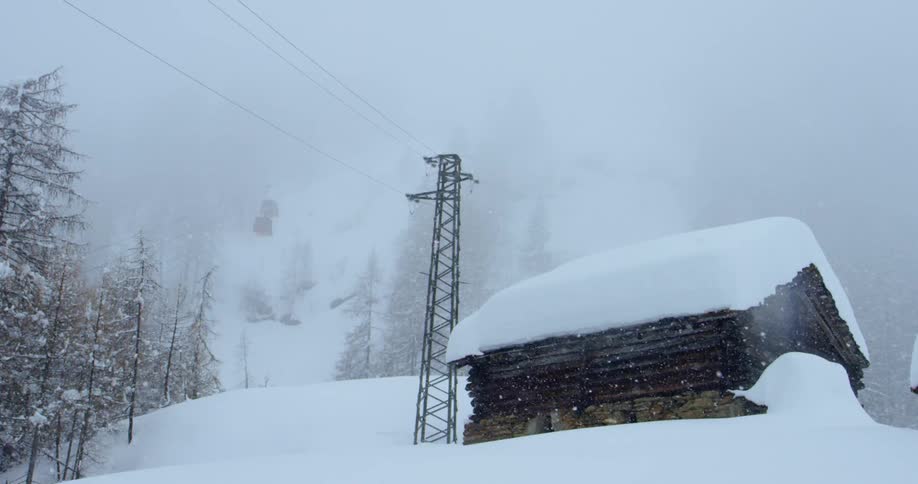 Heavy snow falls from a foggy sky onto a wooden cabin on the outskirts of a forest