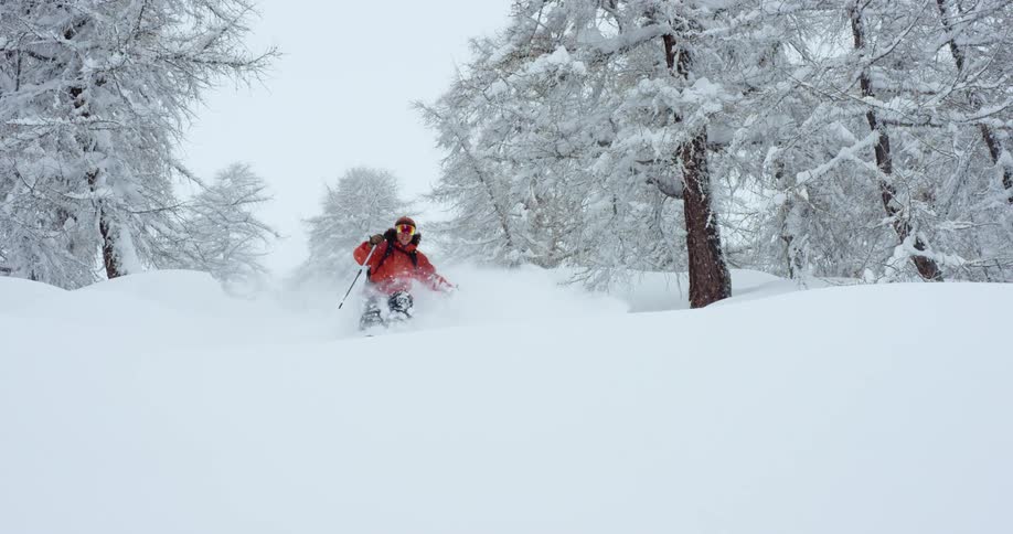 A skier looks into the camera while skiing down a mountain slope as it snows on a winter day