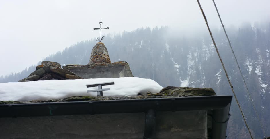 A stone building with a cross with a hill with snow the sky and clouds in the background