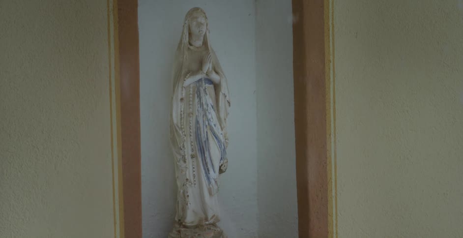 A statue of the blessed virgin mary praying with a rosary with snow falling in the foreground