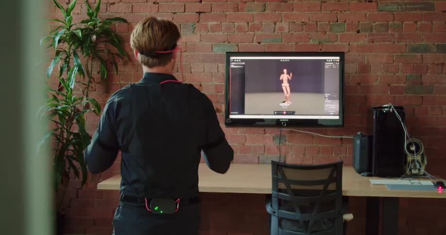 A man wearing motion capture equipment performs movements and watches as they are replicated on a computer monitor