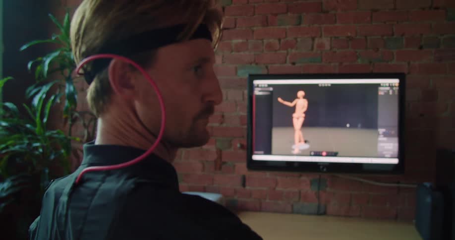 A man with a virtual reality suit moving a figure on a screen next to a table and a plant