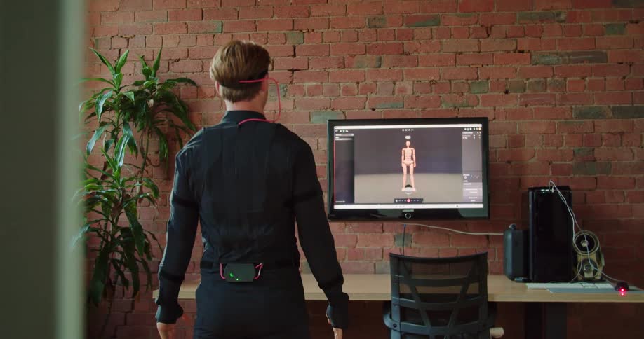 A man stands in front of a monitor using a motion capture suit