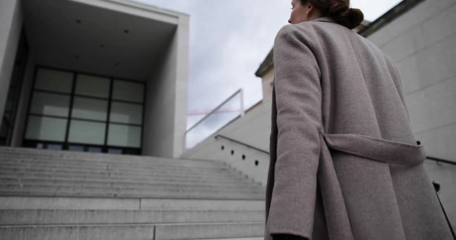 A woman with a coat walking up stairs towards a building with the sky and clouds in the background
