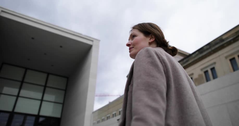 A woman in a longcoat walks up the stone staircase in front of a building on a cloudy day