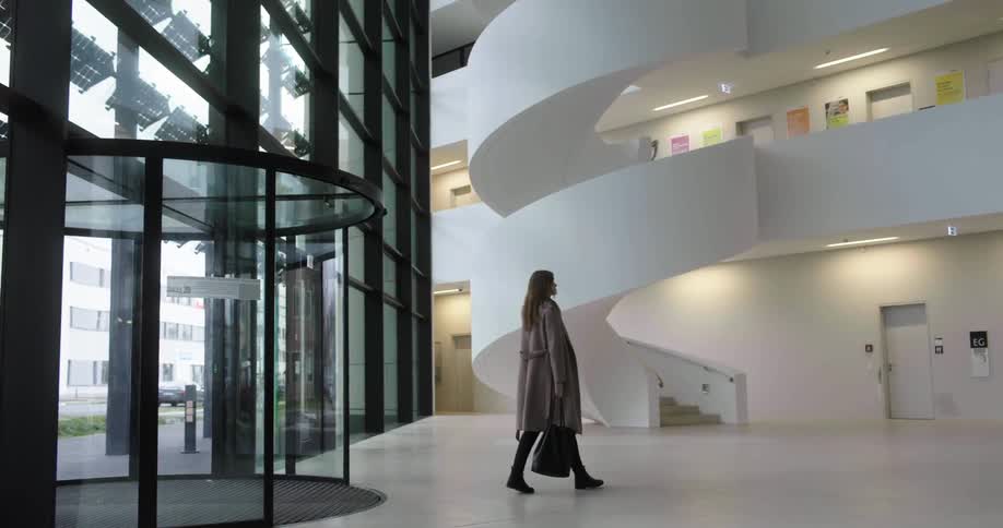 A woman walks into a large modern building with curved white staircases and stops to look up and around