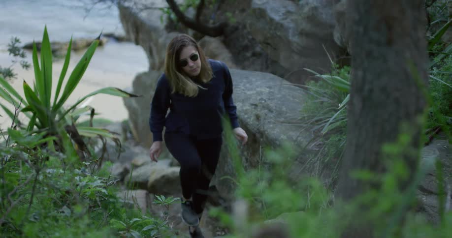 A woman wearing sunglasses hikes up a rocky slope away from a beach