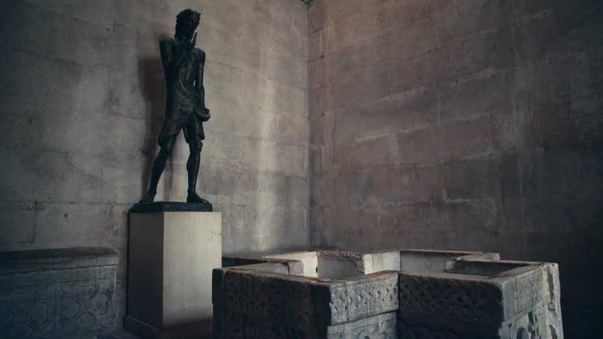 Bronze statue stands on pedestal in marble tiled room near ornate cross shaped basin