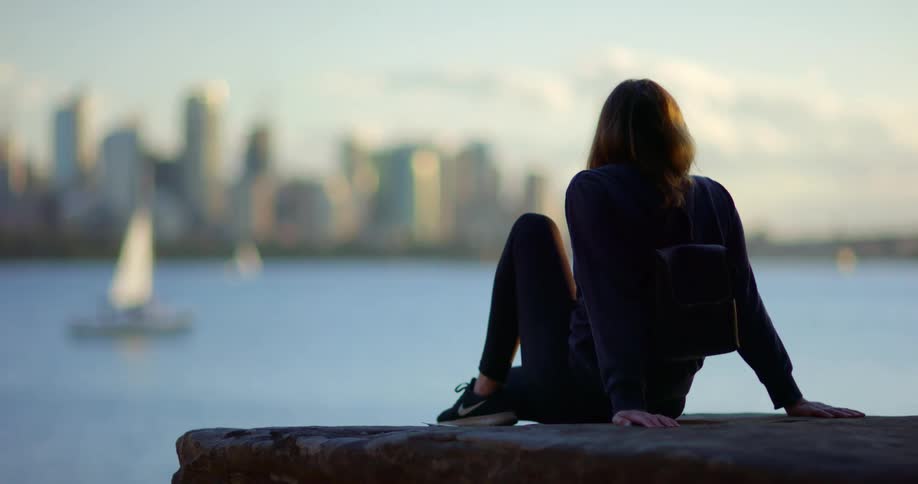 Woman sits on flat stone looking toward city skyline past sailboats on the water
