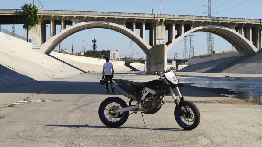 A person holding a helmet with a motorcycle in the foreground and a bridge and the sky in the background