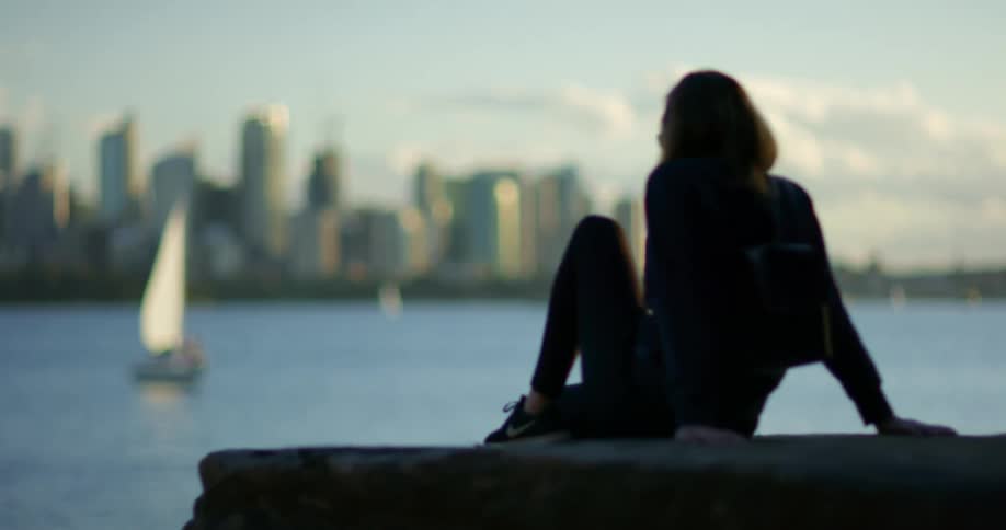 A woman looks out over the bay of a metropolis while sitting on a large stone on the shore