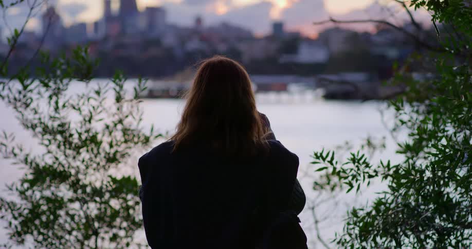 Woman wearing oversized coat looks over water toward city skyline under a red tinted sky