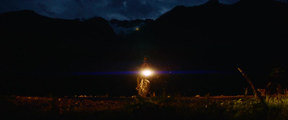 A person sitting on a motorcycle with headlights with a mountain range the sky and clouds in the background