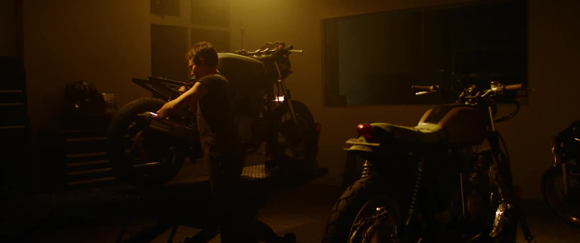 A white boy in a shirt sleeved shirt is standing in a shop filled with motorcycles working on one in the dimly lit room