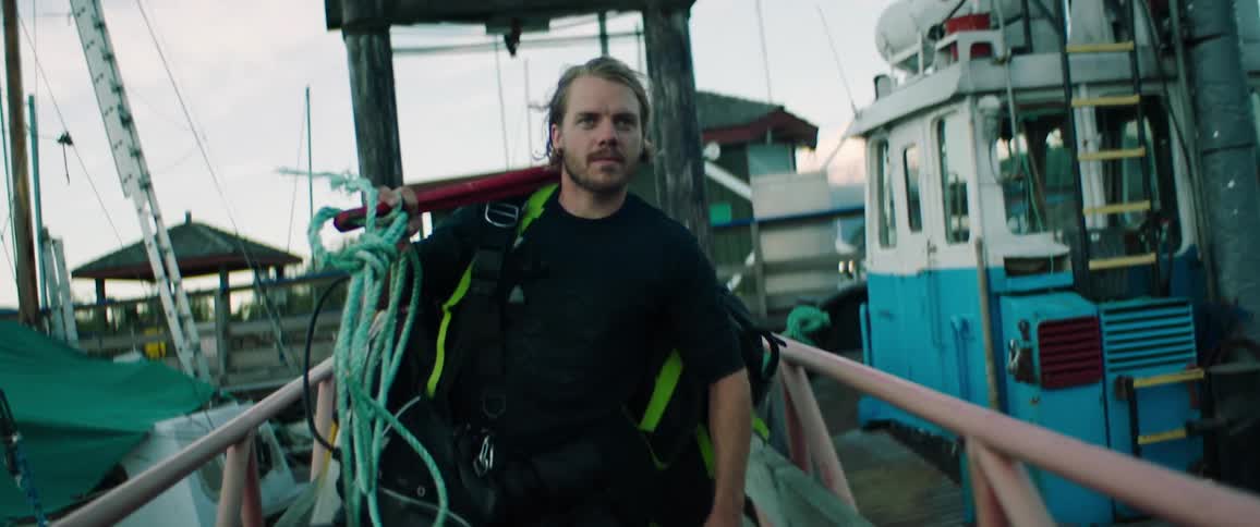 A man carrying scuba equipment walks down a dock past boats