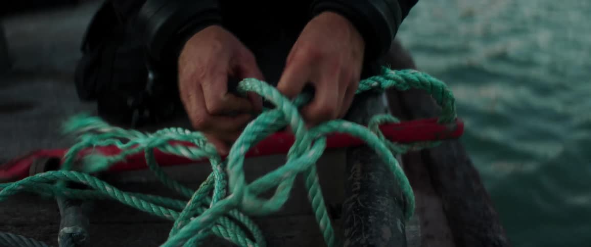 Man in boat ties knot in rope as water ripples between boat and waterfront
