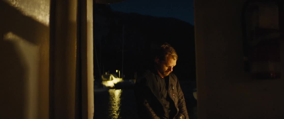 Man works on air tank as light reflects on water from boat near mountain at night